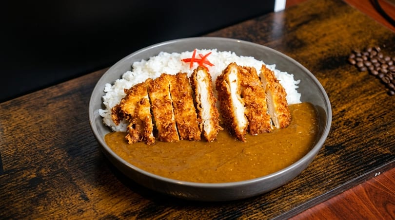 Crispy chicken katsu curry with white rice and red garnish in a gray bowl on a wooden table.